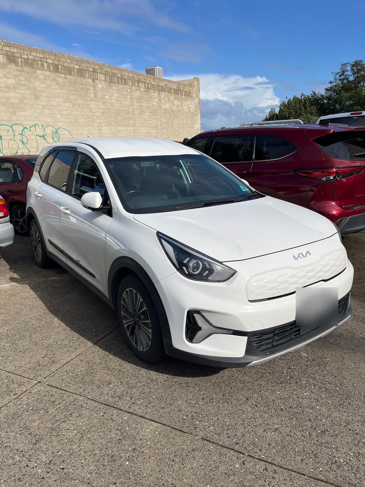 A white Kia SUV parked beside a brick wall, with its number plate blurred